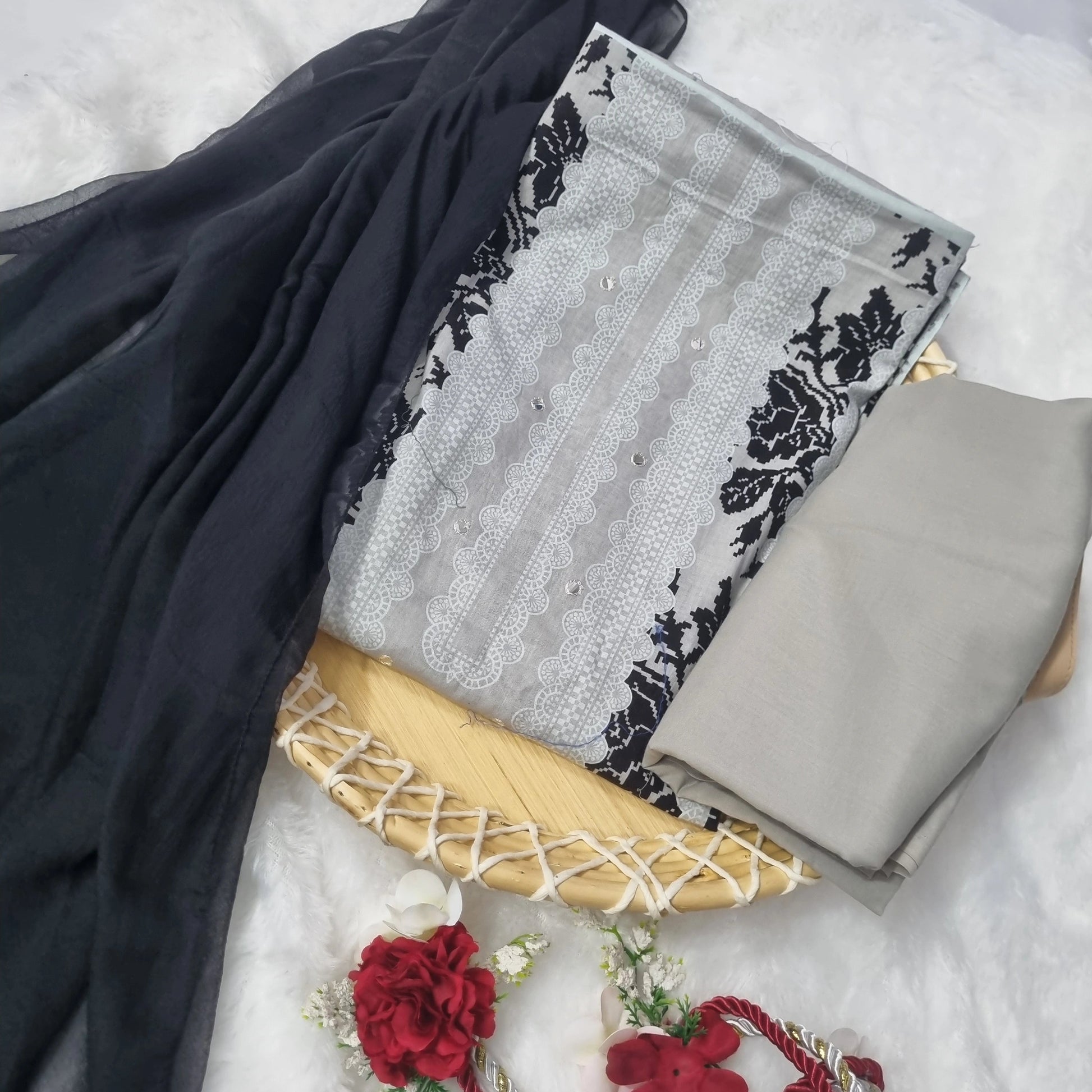 A folded traditional Indian salwar suit set displayed on a white fabric background, featuring a light gray and white embroidered fabric with black floral borders, a solid gray bottom piece, a black dupatta, and decorative red flowers with a red-and-white cord accessory.