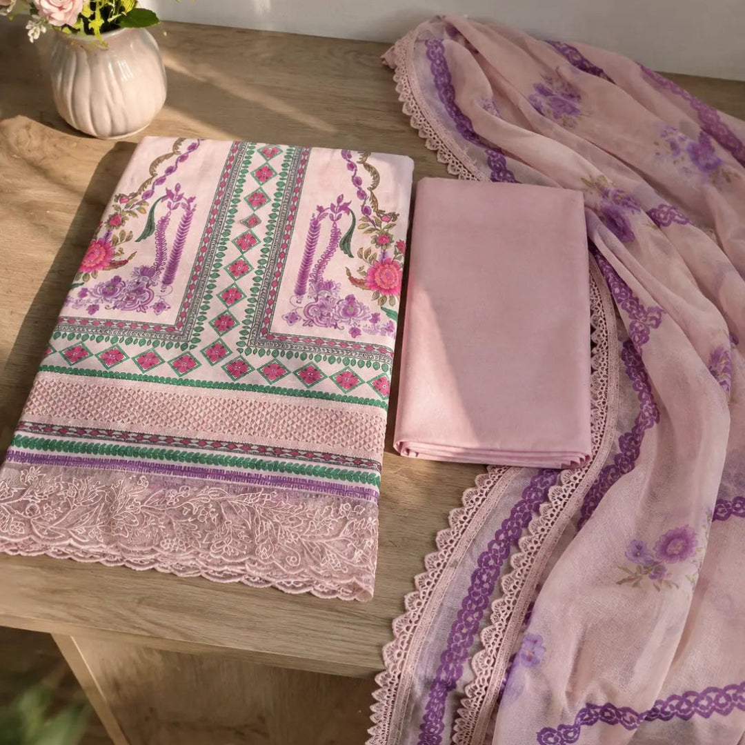 A folded pink fabric set with a patterned textile featuring peacock motifs and geometric borders, alongside a solid pink folded piece and a sheer pink dupatta with purple floral prints and lace trim, arranged on a wooden table with flowers in the background.