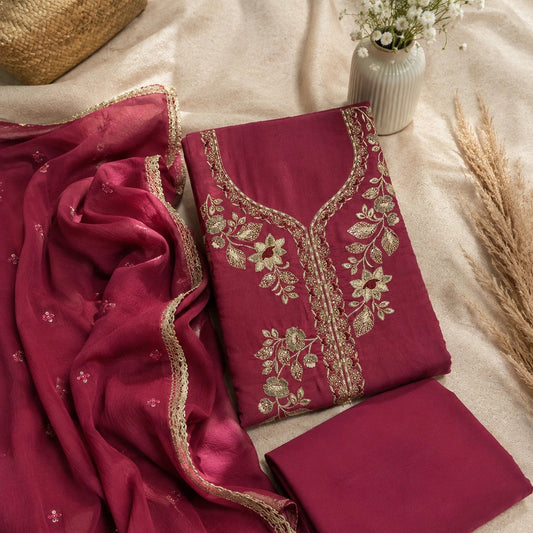 A folded deep magenta salwar suit set with matching dupatta, featuring intricate gold floral embroidery on the neckline and border details, styled on a beige fabric background with decorative dried grass and a small vase of flowers.