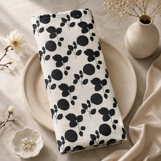 A folded white fabric item, likely a blanket or throw, featuring a bold black floral and leaf pattern, styled on a ceramic plate with neutral props.