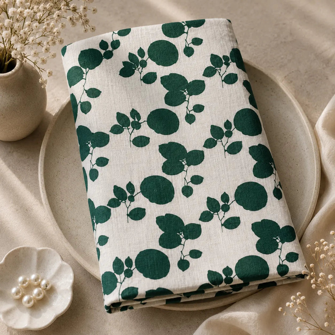 A folded white fabric item, likely a muslin cloth or napkin, featuring a dark green botanical pattern of leaves and circles, styled on a beige ceramic plate with dried flowers and pearl beads.