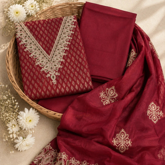 A folded and draped deep red or maroon traditional fabric set with beige embroidered borders and motifs, arranged in a wicker basket with white flowers on a beige fabric background.