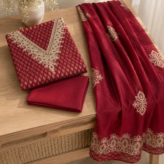 A folded deep red traditional fabric set with cream embroidered borders and a matching draped dupatta, displayed on a wooden table next to a vase of white flowers.