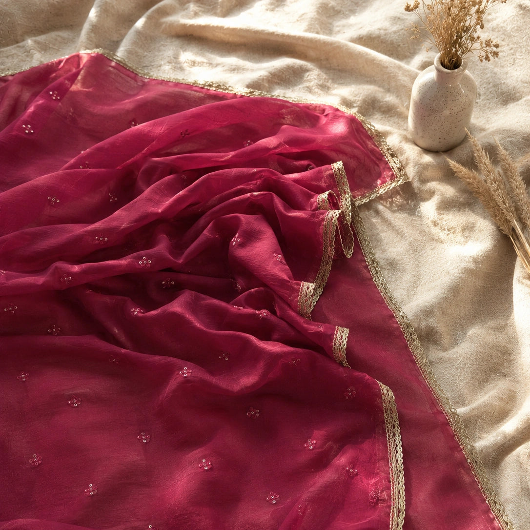 A deep magenta pink dupatta with delicate gold zari embroidery and border trim, draped elegantly on a beige fabric surface next to a small vase with dried flowers.
