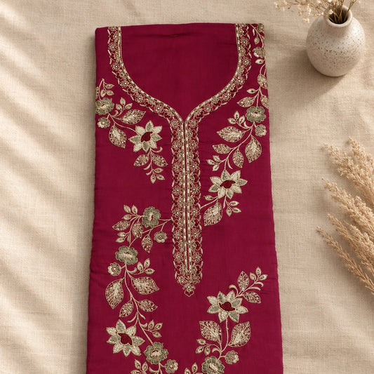 A folded deep magenta pink fabric garment, likely a kurta or tunic, featuring intricate gold and silver floral embroidery along the neckline and down the center front. The garment is laid flat on a beige linen fabric background with dried decorative grass and a small vase in the corners.