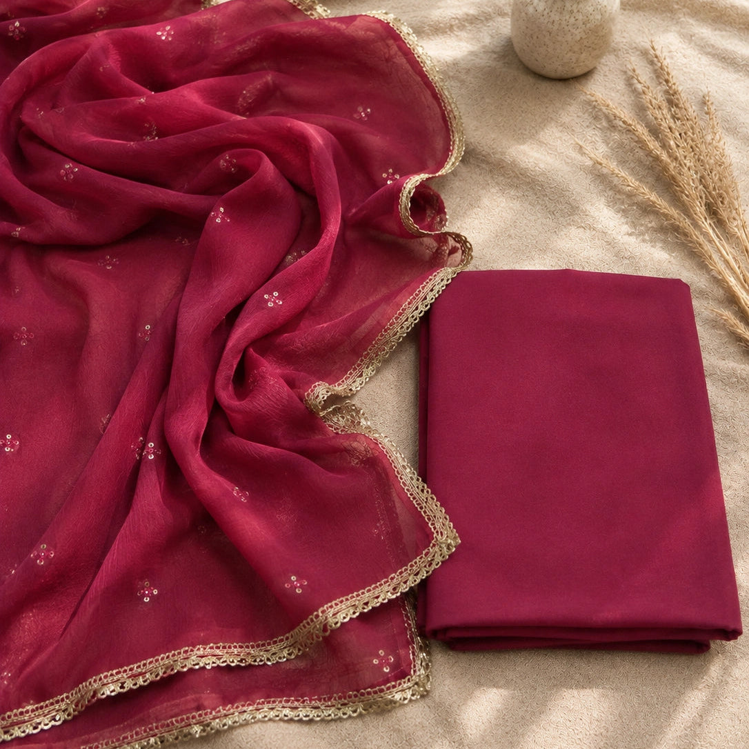 A folded and draped magenta dupatta with gold lace borders and subtle floral embroidery, displayed on a beige fabric background next to a folded matching fabric piece, with dried decorative grass and a small vase in the background.