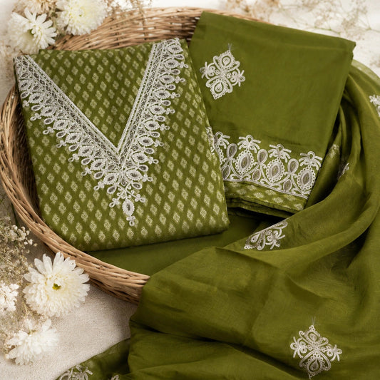 An olive green ethnic salwar kameez fabric set with white embroidery, displayed in a wicker basket with white flowers on a neutral background.