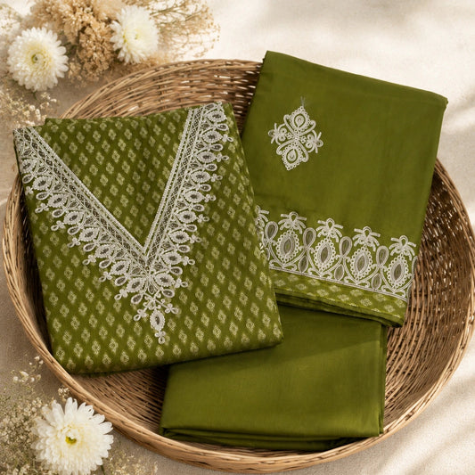 An olive green fabric set with white embroidery and lace borders, neatly folded and displayed inside a round woven wicker basket on a beige fabric surface with decorative white flowers.