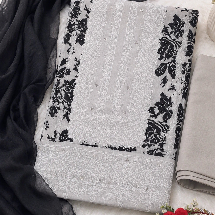 A folded light gray and black embroidered fabric set with a matching sheer black dupatta, arranged on a white textured surface with decorative flowers and a red rose accessory.