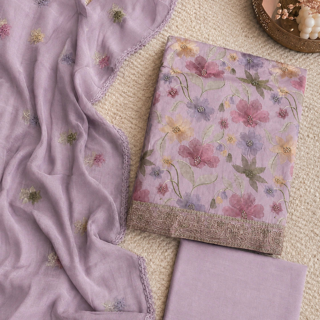 A folded and draped floral fabric set in soft lavender, including a patterned cloth with embroidered flowers, a matching solid fabric piece, and a sheer embroidered scarf, arranged on a textured beige surface.