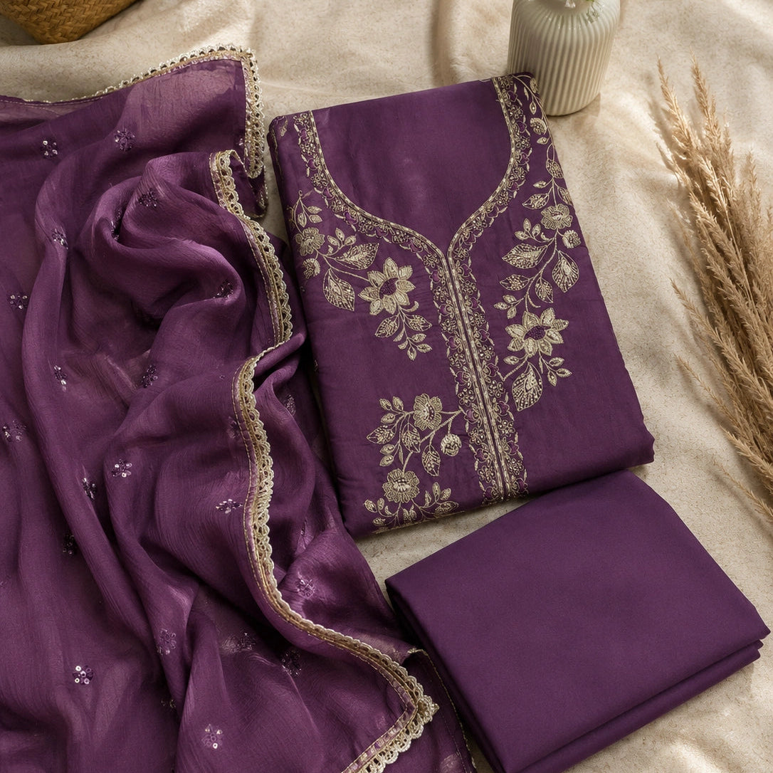 A folded purple traditional outfit set with matching dupatta, laid out on a beige fabric surface with decorative dried grass and a small vase of flowers. The set includes a purple kurta/tunic with beige floral embroidery and a matching folded fabric piece.