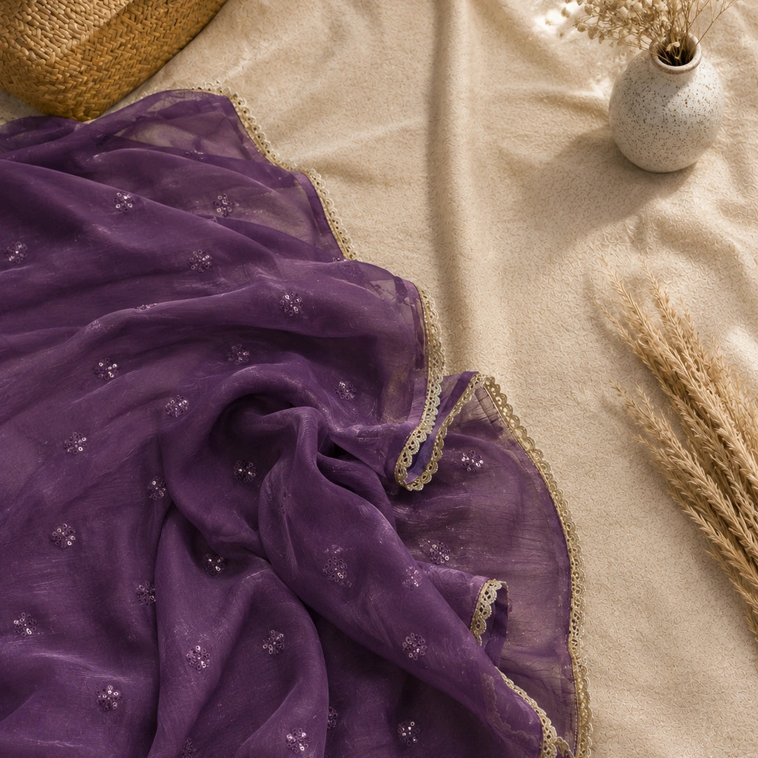 A draped purple sheer fabric with small embroidered motifs and a beige lace border, styled on a beige fabric surface with decorative dried grass and a small vase.