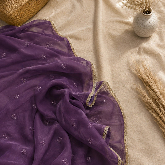 A draped purple sheer fabric with small embroidered motifs and a beige lace border, styled on a beige fabric surface with decorative dried grass and a small vase.