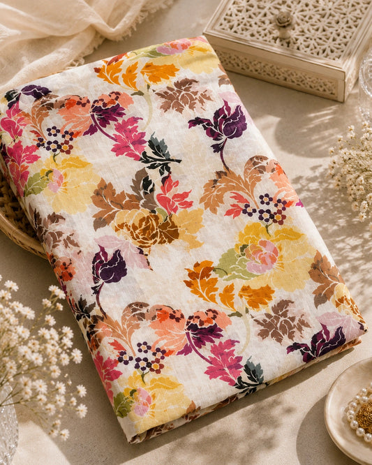 A folded cotton fabric with a vibrant floral print in shades of pink, orange, yellow, purple, and brown on a cream background, styled on a neutral beige surface with decorative props like a lace box, dried flowers, and a pearl dish.