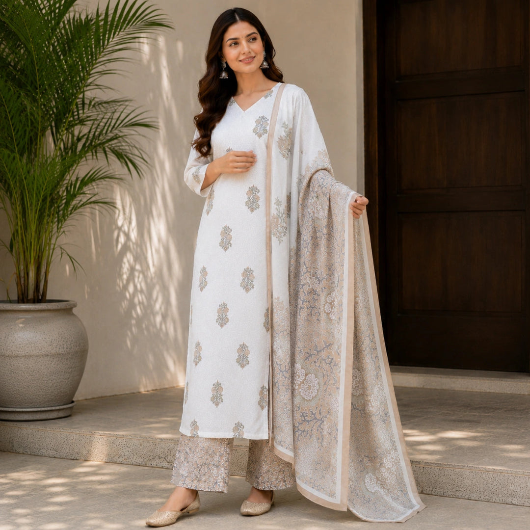A woman models a white three-piece traditional outfit with beige floral embroidery and a matching sheer dupatta, standing in front of a neutral wall and dark wooden door.