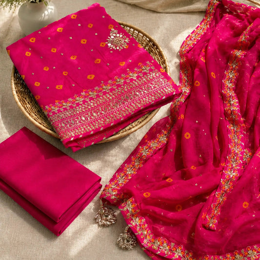 A folded and draped bright pink traditional fabric set with embroidered floral borders, displayed on a neutral fabric surface with decorative dried grasses and a small vase of white flowers.