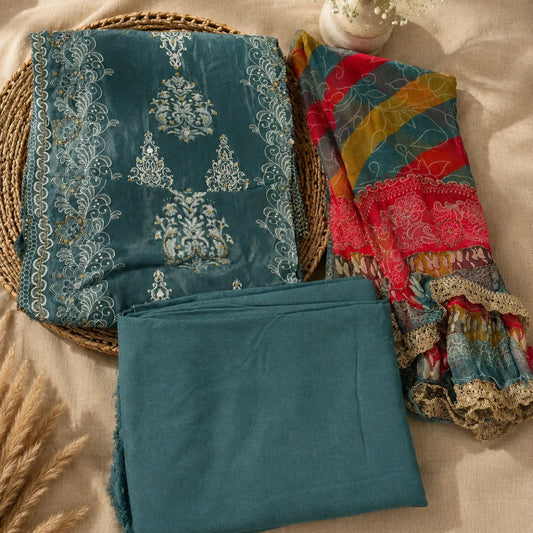 A folded teal fabric set with ornate white embroidery and a multicolored patterned scarf, styled on a beige fabric background with a woven tray, dried grass, and a small vase of flowers.