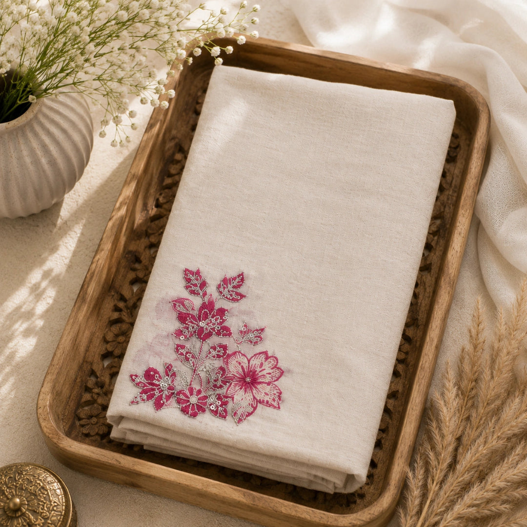 A folded cream-colored linen cloth with a pink floral embroidery design in the lower-left corner, presented in a rustic wooden tray with decorative dried grass and white flowers.