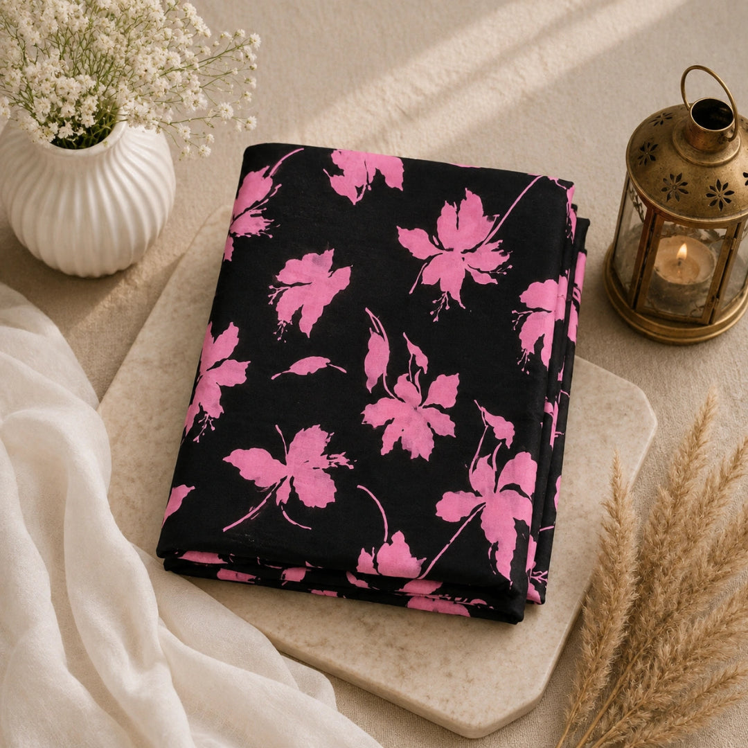 A folded black fabric item with a bright pink floral print sits on a light stone board, styled with a white vase of baby's breath, a brass lantern, and dried grasses.
