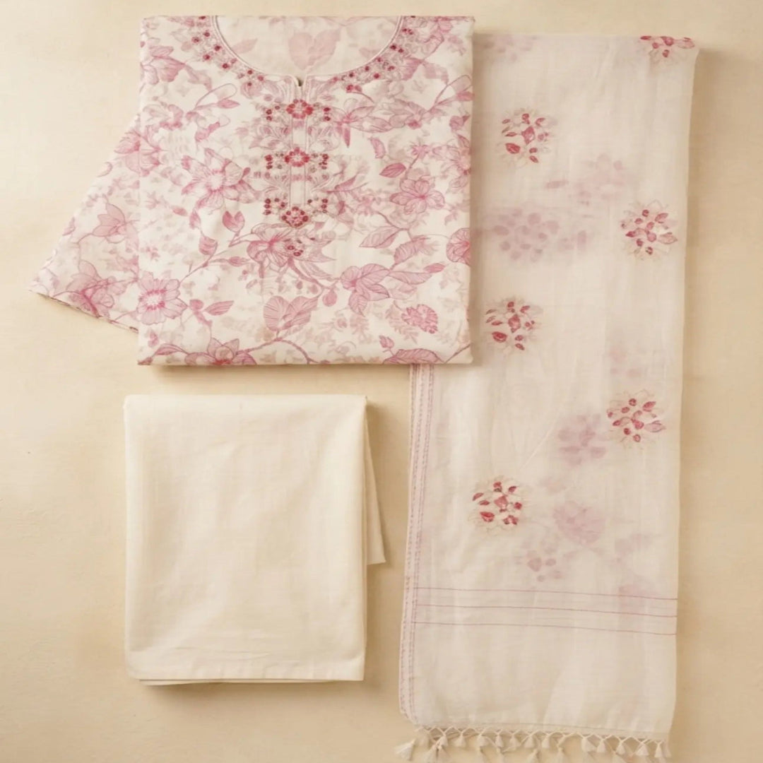 A folded cream-colored cotton fabric set with a pink floral print and a matching cream dupatta featuring pink floral motifs and tassels, laid out on a beige background.